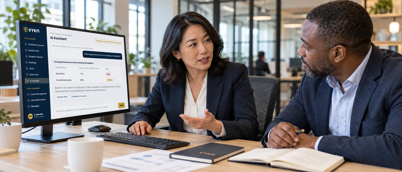 A compliance officer in conversation with a colleague at a desk, with the VYKN AI Assistant open on the monitor.
